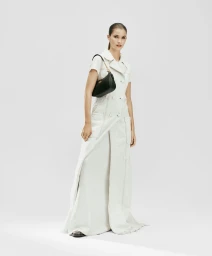 Model River Klein in a full-length white coat dress carrying a black Coach shoulder bag against a white seamless backdrop.