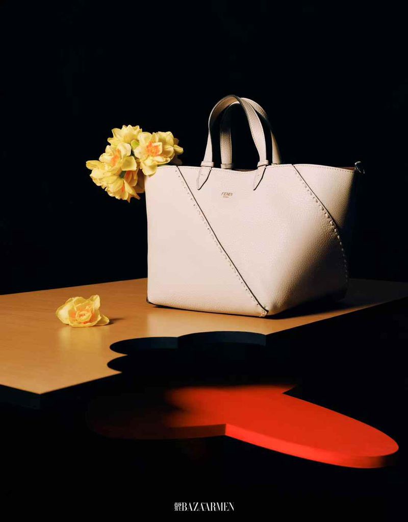 Elegant tote bag on a table with yellow flowers against a black background, illuminated by soft light in a studio setting.