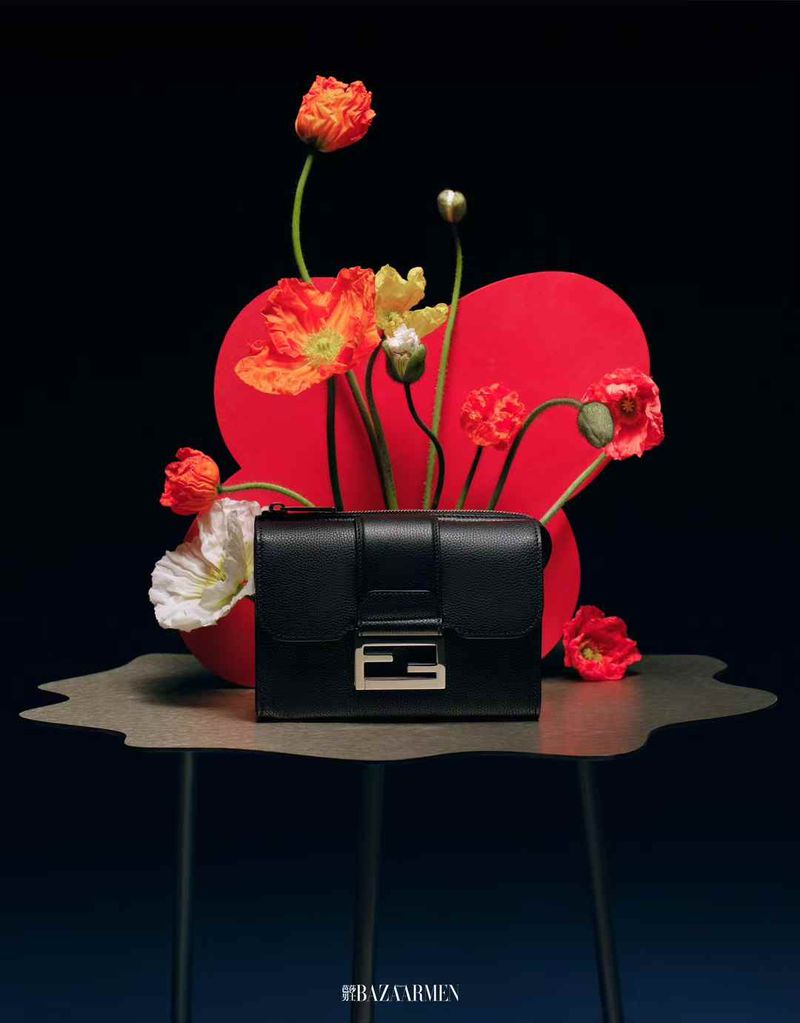Black leather purse with silver hardware on a table surrounded by red, white, and orange flowers against a dark background.