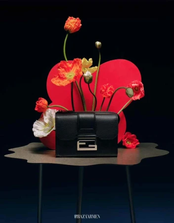 Black leather purse with silver hardware on a table surrounded by red, white, and orange flowers against a dark background.