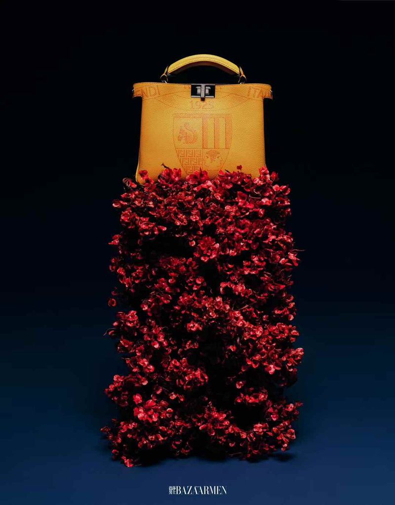 Fendi purse sits on red flower arrangement against a blue backdrop. The bag has a handle, gold accents and the brand name on the front.
