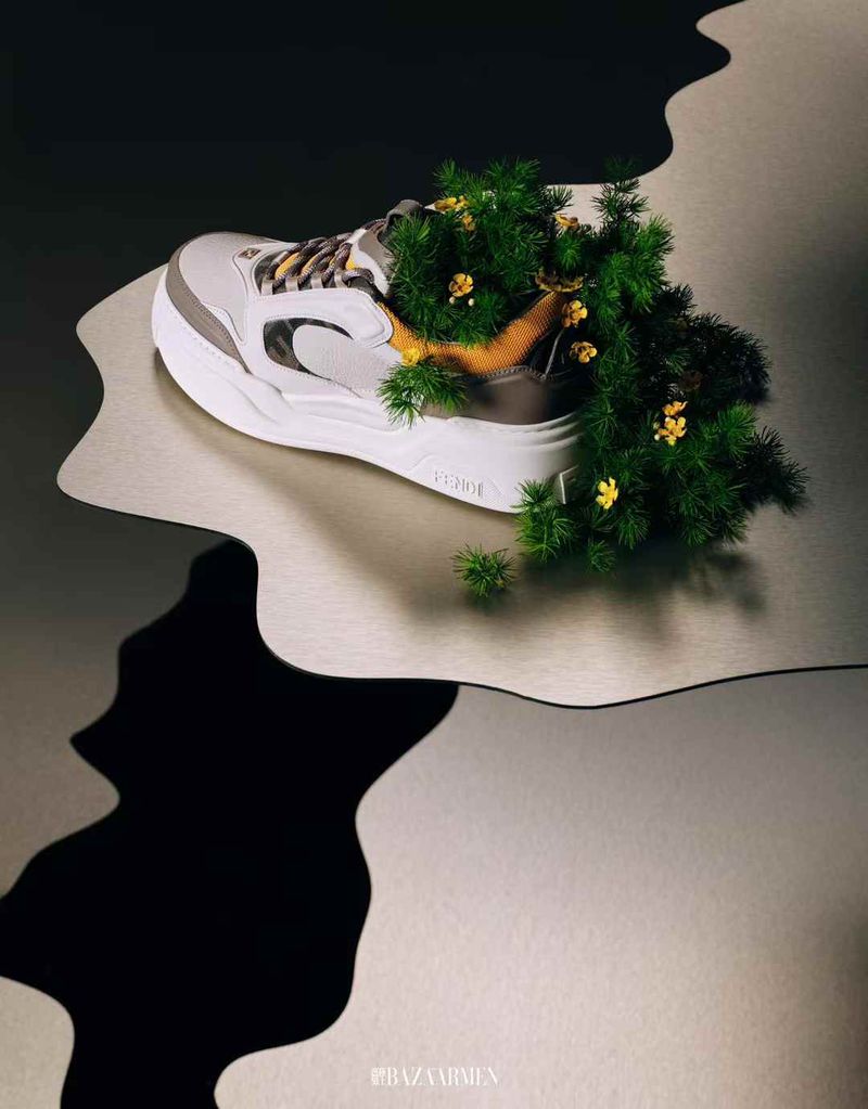 Studio shot of a white and grey athletic shoe filled with green plants and yellow flowers, against a black and silver background.