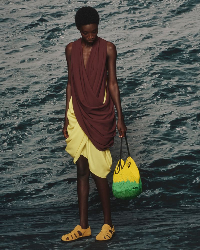 Model wearing a draped burgundy top, pale yellow skirt, and yellow sandals holding a beaded bucket bag against dark ocean water.