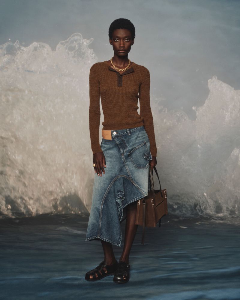 Model posing in a brown knit polo, asymmetrical denim skirt, and leather sandals in front of a crashing ocean wave backdrop.