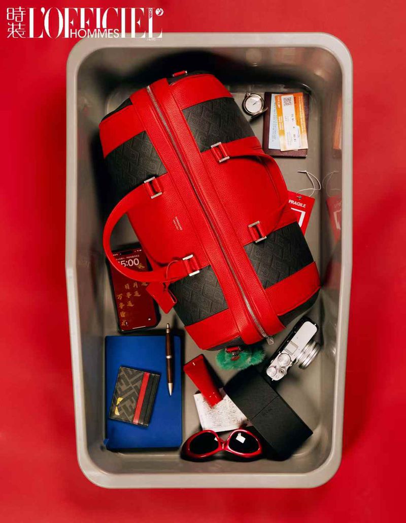 Overhead shot of a red and black travel bag inside a grey security bin with accessories and toiletries on a red background.