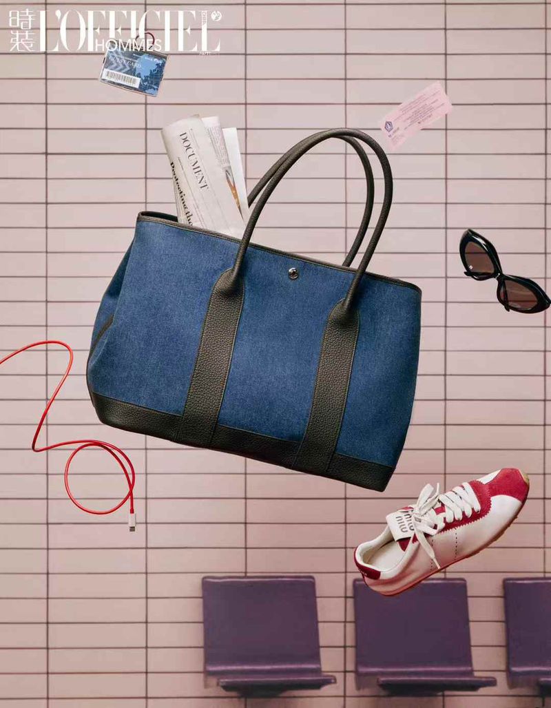 Fashion editorial shot of a denim tote bag with accessories, including a sneaker, sunglasses, a magazine, and a cable, floating mid-air.