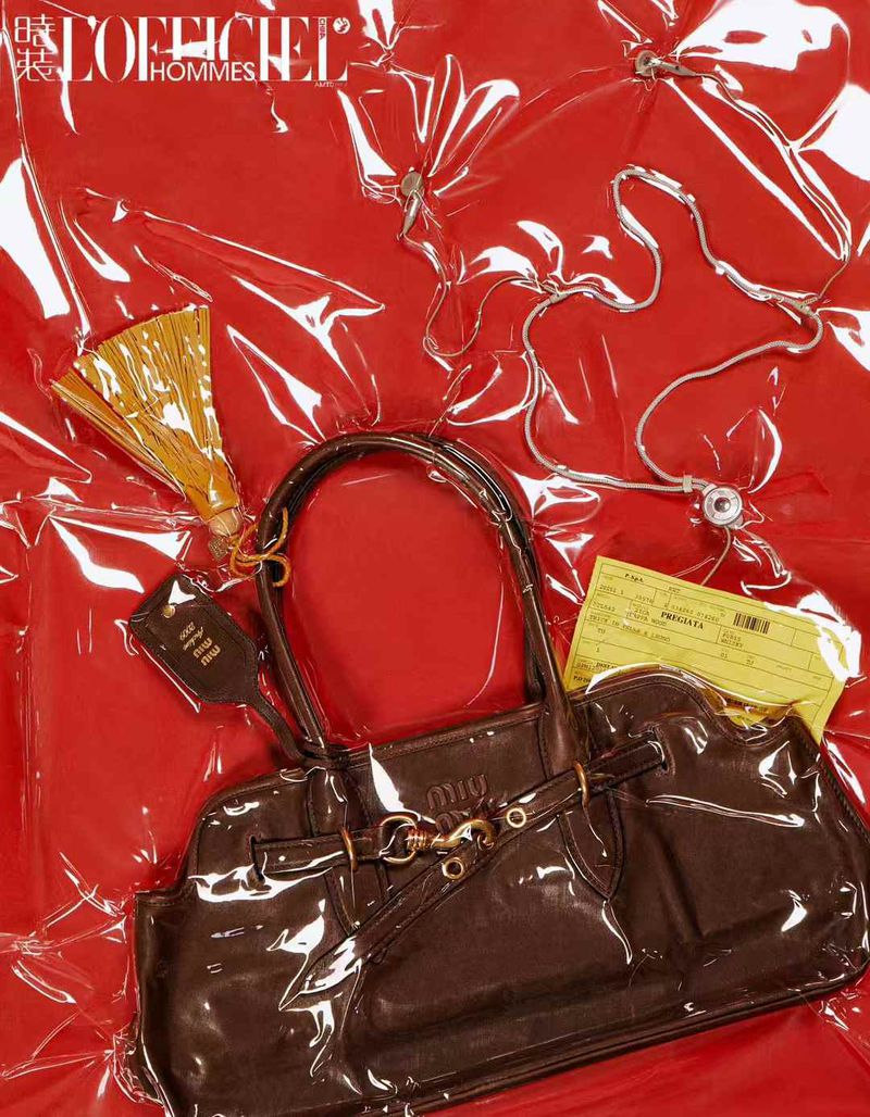 Luxury brown patent leather handbag with gold accents and a tassel, displayed on wrinkled red plastic sheeting, magazine ad.