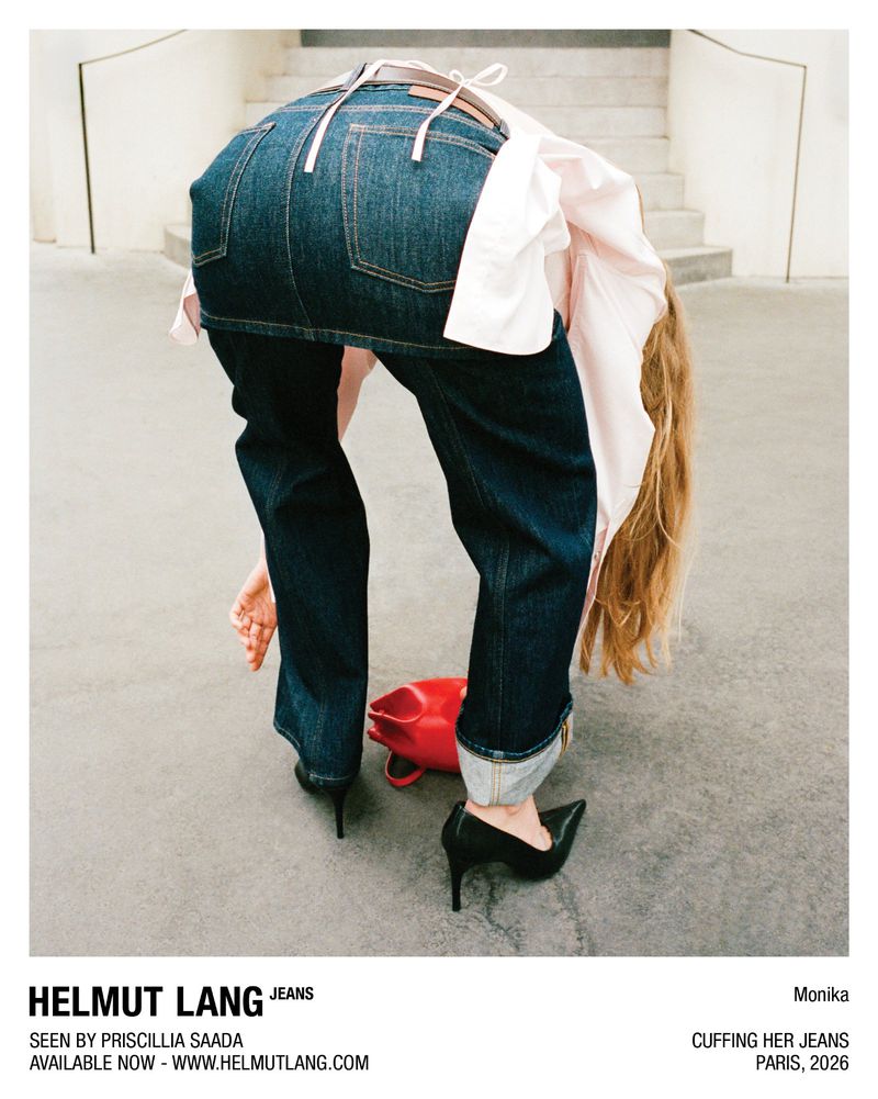 Woman bending forward wearing a denim skirt over cuffed jeans, a pale collared shirt, and black heels on outdoor pavement.