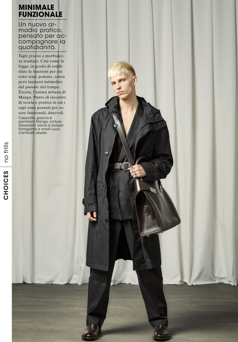 A male model in a black coat and dark trousers holds a brown leather bag against a minimalist background in a fashion editorial.