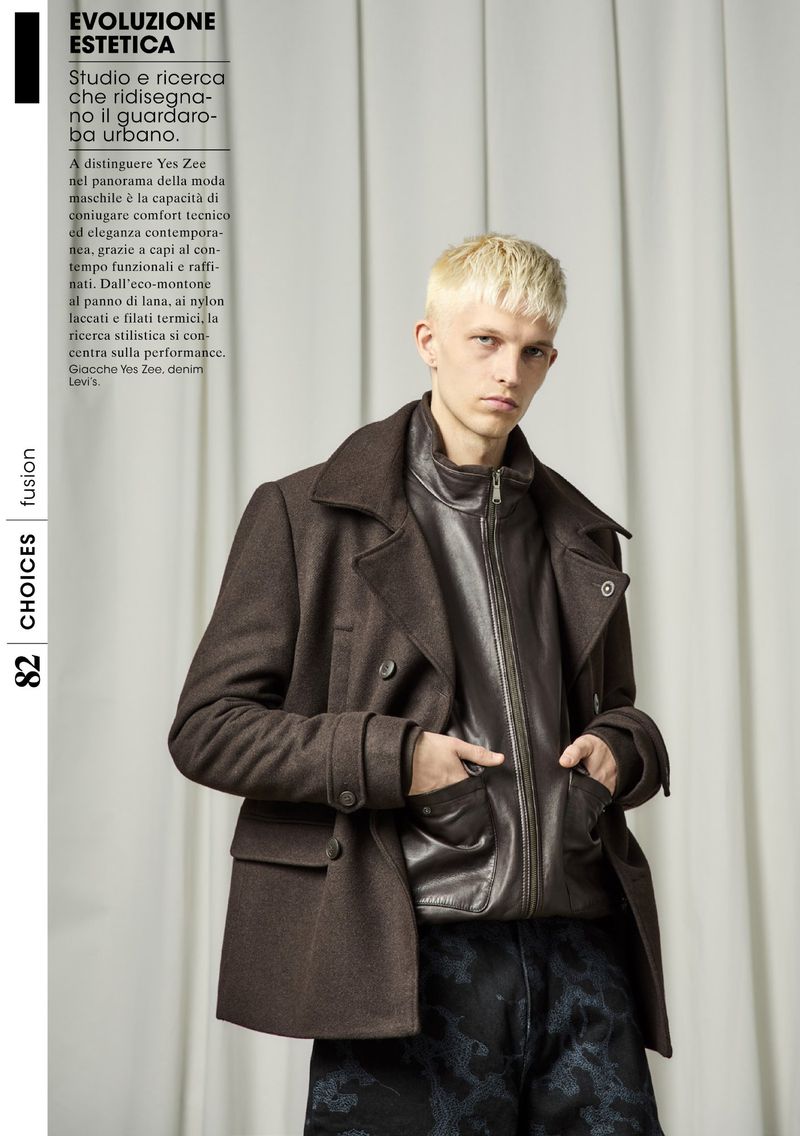 Blonde male model poses in a stylish dark brown peacoat and leather jacket against a neutral curtain backdrop for a fashion editorial.