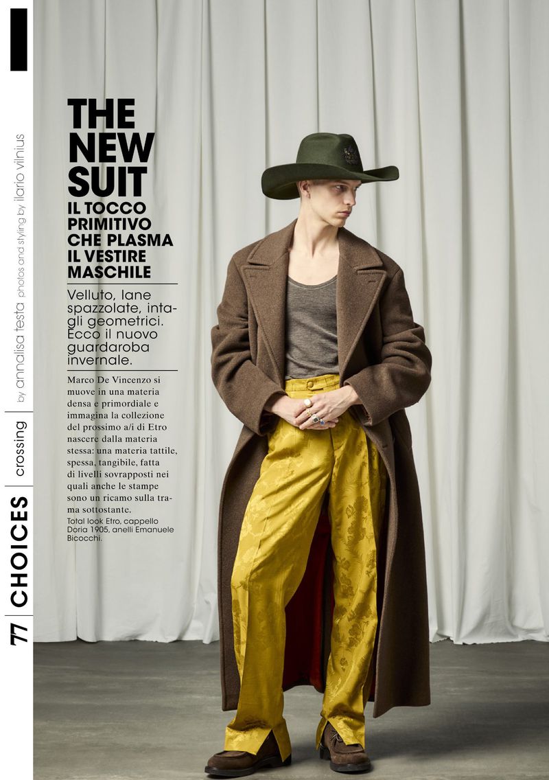Fashion editorial shot of a slender male model wearing a hat, long coat, tank top, patterned trousers, and shoes against a light backdrop.