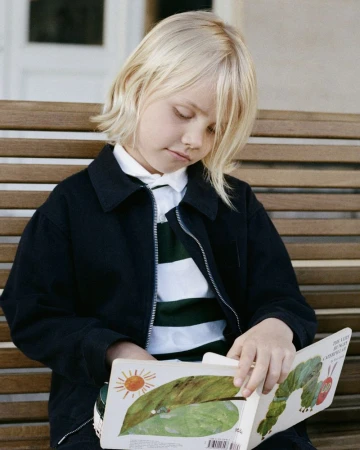 A blonde child sits on a slatted wooden bench reading The Very Hungry Caterpillar wearing a black zip jacket and striped collared shirt.