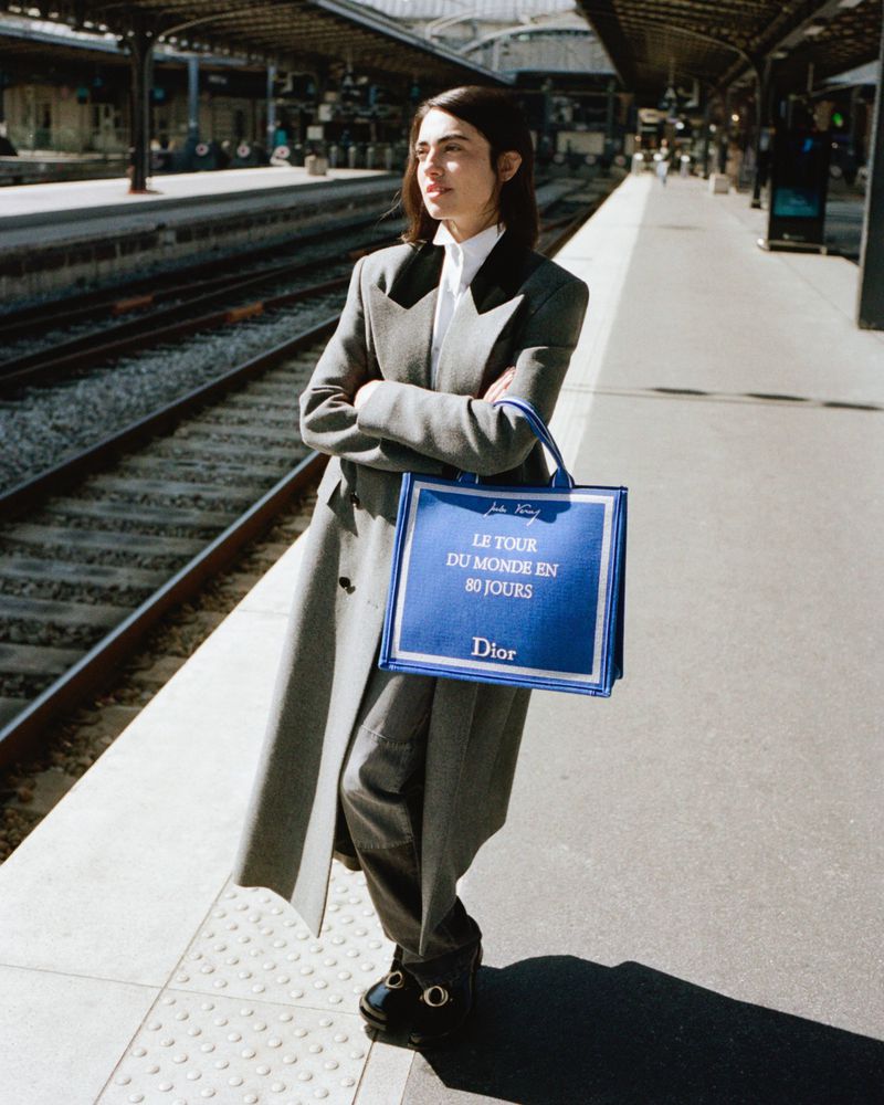 Model wearing a tailored grey coat holding a blue Dior tote bag standing on a sunny railway station platform.