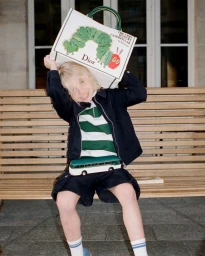 Child sitting on a wooden bench holding a Dior Book Tote featuring The Very Hungry Caterpillar artwork over their head.