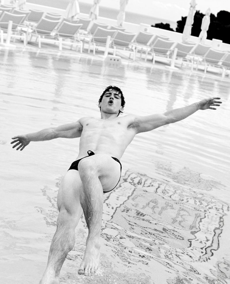 Athletic man with open arms descends toward pool. Swimsuit and muscular build visible. White lounge chairs in blurred background.