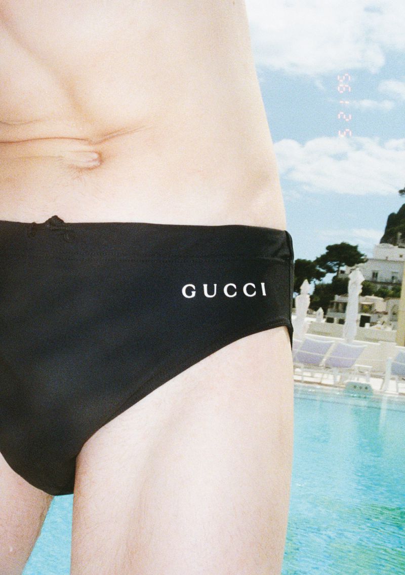 Midsection of a man wearing black Gucci swimwear, posing near a swimming pool with a resort and cloudy sky in the background.
