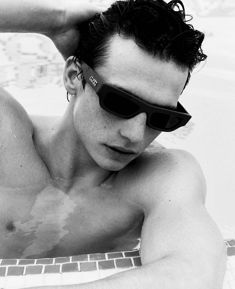 Monochromatic portrait of a shirtless man wearing sunglasses, posing in a pool with patterned tiles, wet hair, and a contemplative expression.