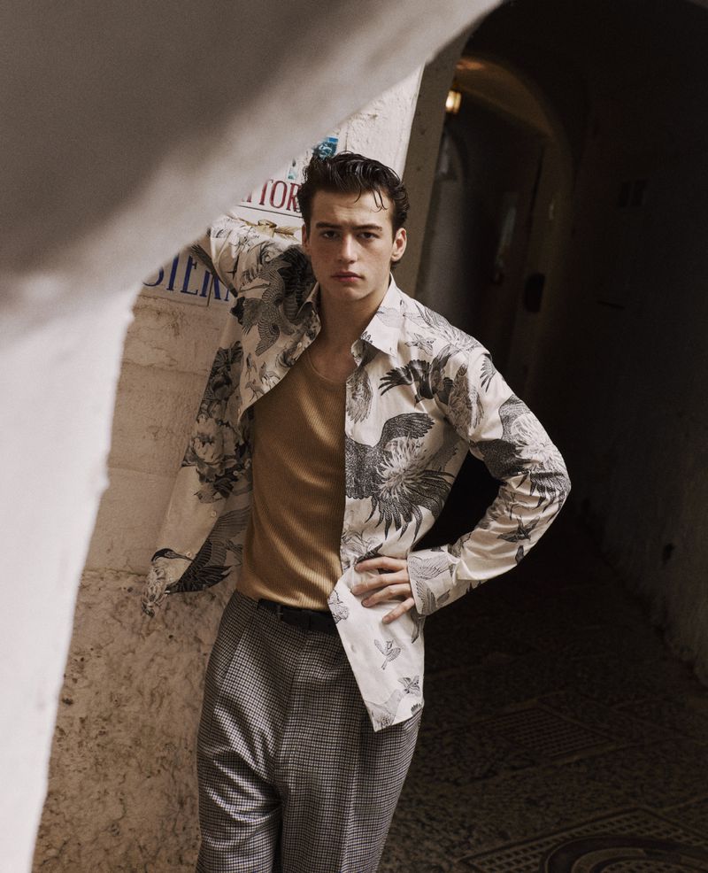Young man poses indoors in a tunnel or arched alcove, wearing a patterned shirt and pants. Fashion shot. Light complexion.