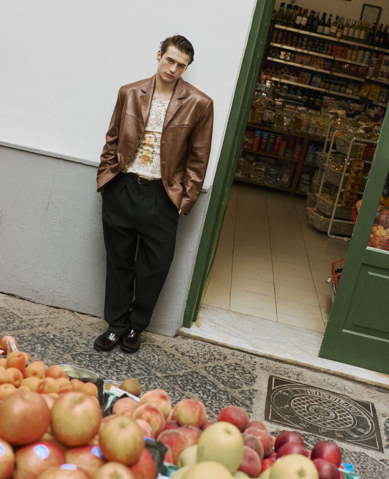 Fashionable man in brown leather jacket poses next to a fruit stand outside a grocery store, creating a unique street style portrait.