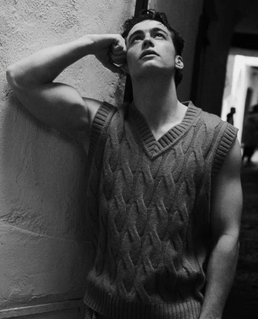 Monochrome portrait of a muscular man in a sleeveless knitted sweater, looking up while leaning against a wall in a dark indoor setting.