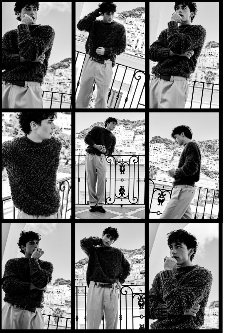 Black and white fashion shot of a young adult man wearing a wool sweater, standing on a balcony with a seaside town in the distance.