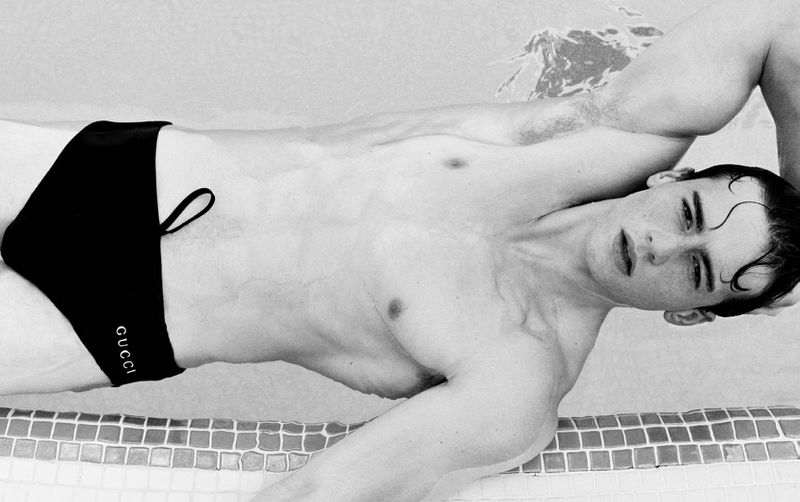 Monochrome portrait of a young adult man in a black Gucci swimsuit, lying in water with a tiled edge in the background.