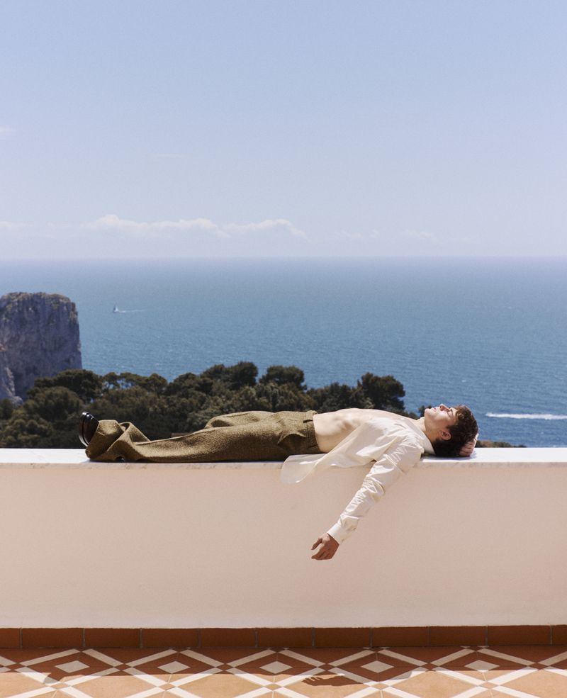 Adult man lying on a white wall, wearing a white shirt and olive-green pants, relaxing on a building terrace with a sea background on a sunny day.
