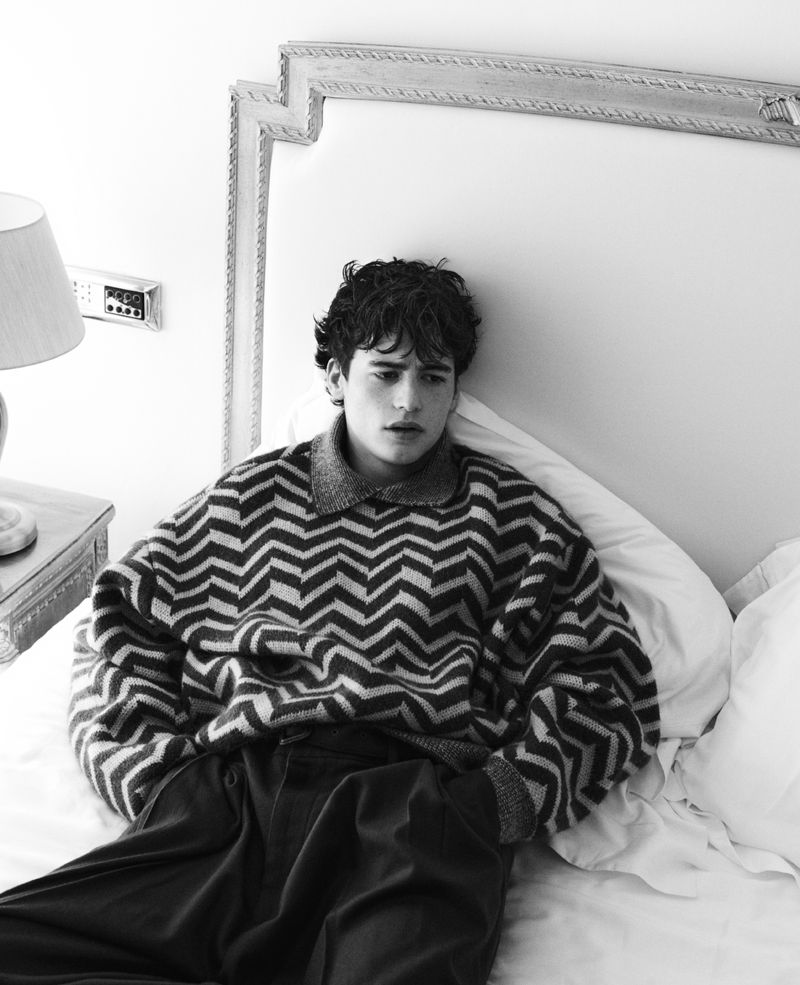 Young man reclining on a white bed wearing an oversized chevron patterned knit sweater and dark trousers.