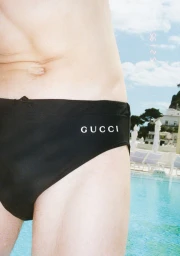 Cropped view of a man wearing black Gucci swim briefs standing beside a bright turquoise outdoor swimming pool.