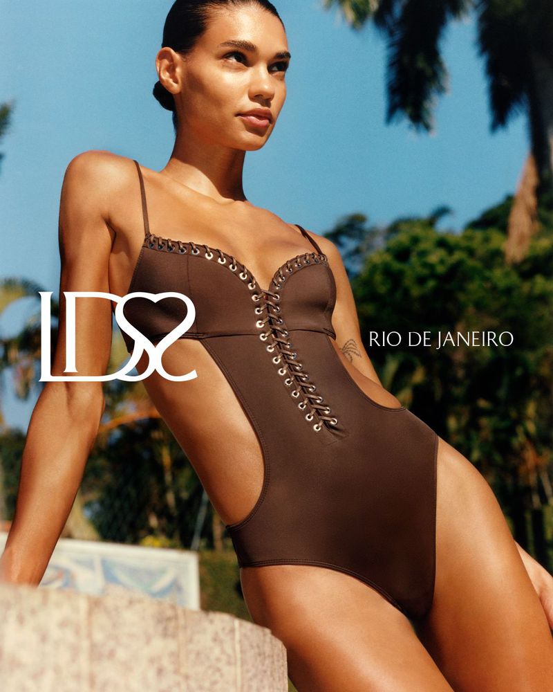 Model Barbara Valente wearing a chocolate brown lace-up cutout swimsuit outdoors against palm trees and blue sky.