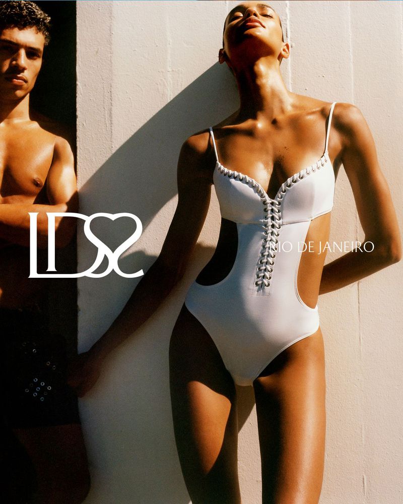 Model Barbara Valente wearing a white lace-up one-piece cutout swimsuit against a sunlit wall next to a shirtless man.