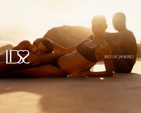 Model Barbara Valente wearing a black lace-up bikini reclining on sand alongside two models at golden hour in Rio de Janeiro.