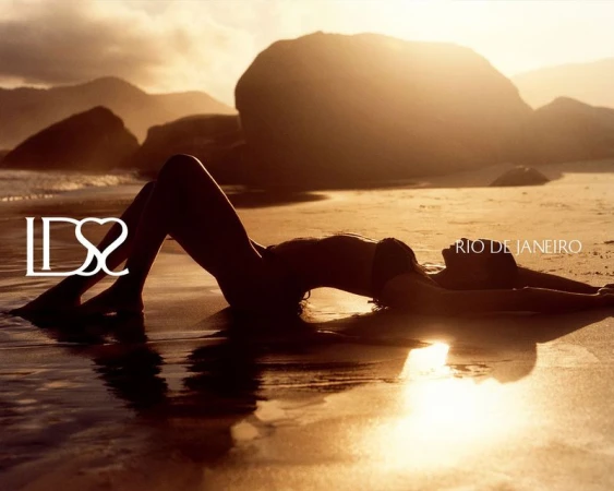 Silhouette of a woman in a bikini reclining on wet sand at sunset against coastal rocks in Rio de Janeiro.