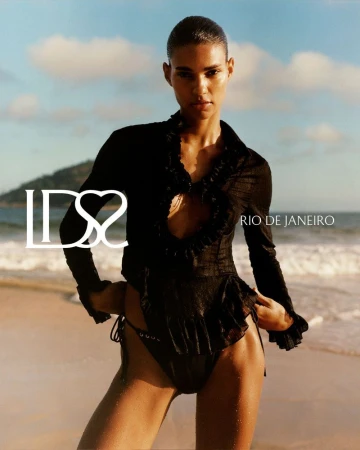 Model Barbara Valente posing in a black ruffled long-sleeve top and bikini bottoms on a sandy beach in Rio de Janeiro.