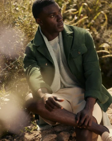 An adult male with dark skin wearing a green jacket and tan shorts is sitting outdoors surrounded by grass and other foliage.