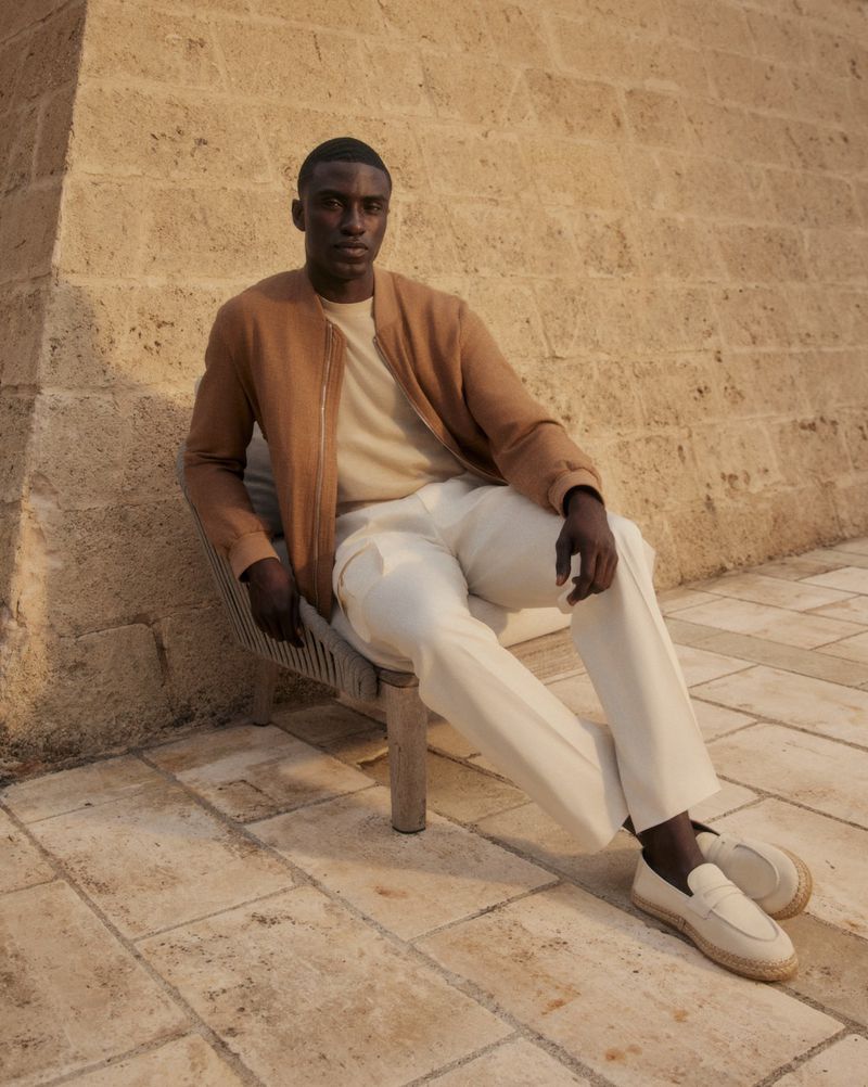 A dark-skinned man sits on a chair in a tan bomber jacket and off-white pants in an outdoor setting against a brick wall.