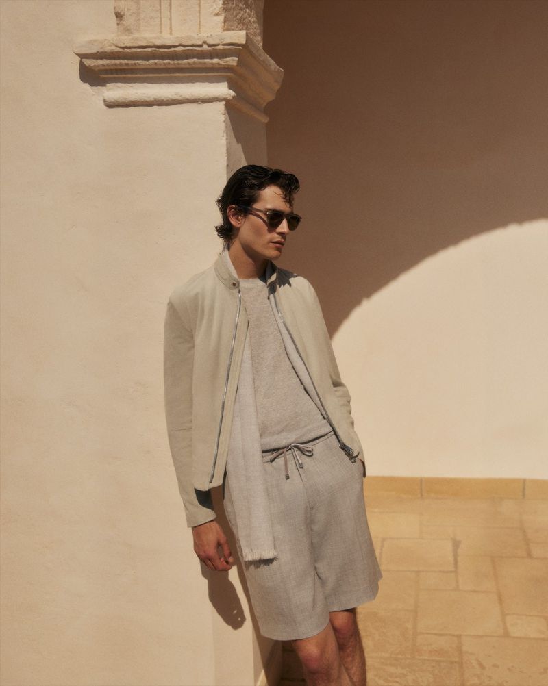 A young adult male in casual resort wear poses against an architectural wall in the sunlight, wearing sunglasses and a jacket.