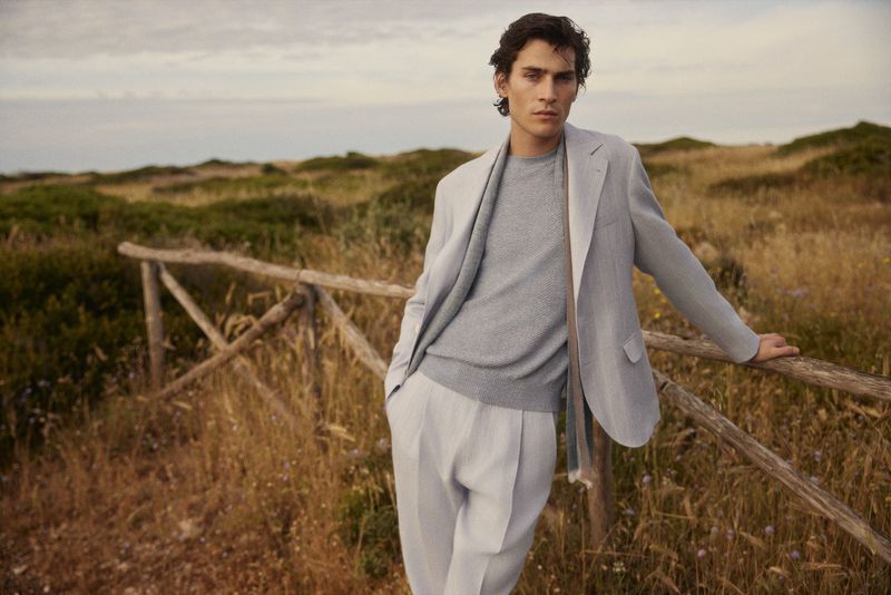 Adult male model poses leaning on a wooden fence in an open field wearing a suit, sweater, and scarf under a cloudy sky.