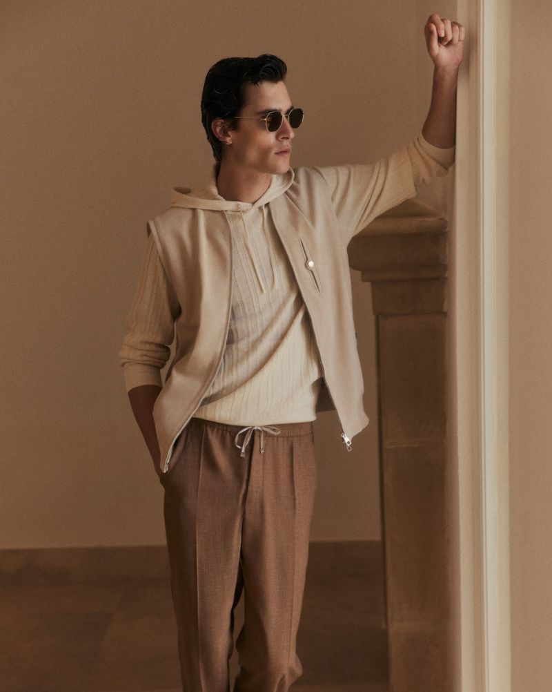 A stylish adult male poses indoors with a relaxed expression, wearing a hooded vest and sunglasses, leaning against a wall in soft lighting.