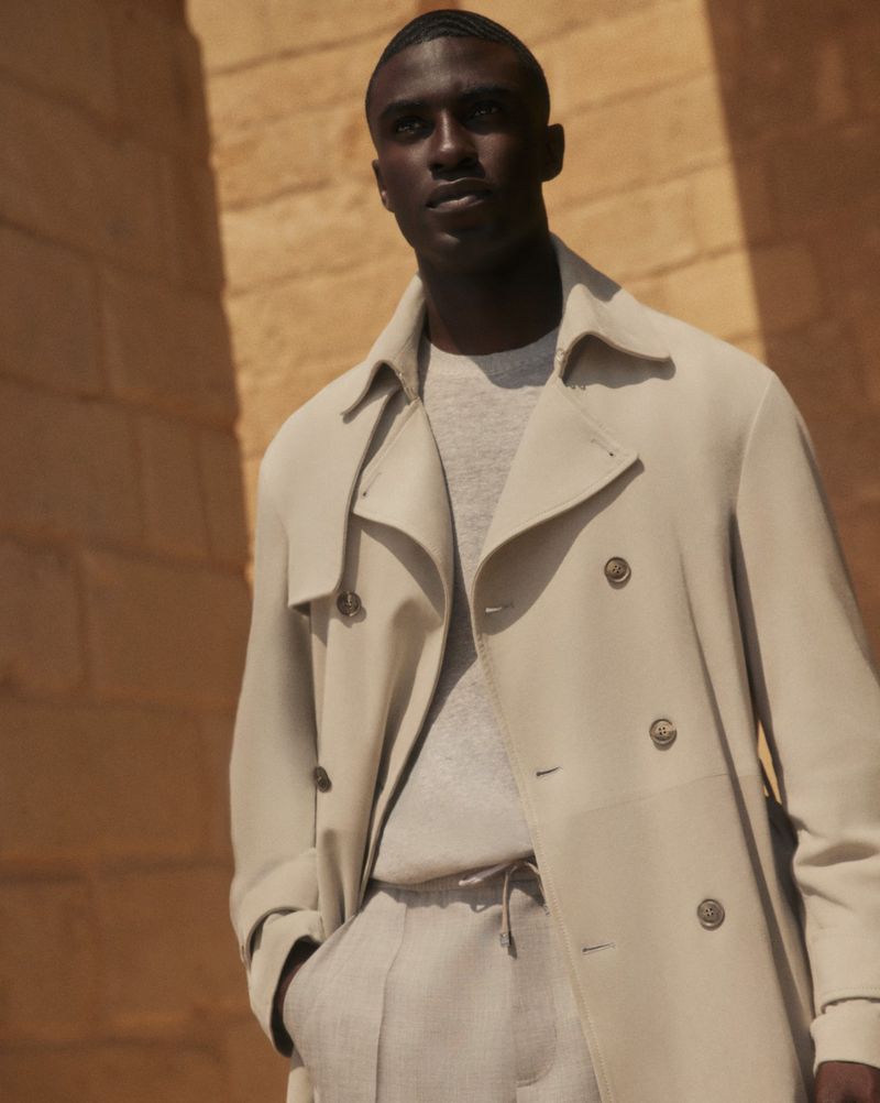 A stylish black man in a beige trench coat poses against a textured stone wall, showcasing fashion and urban style with neutral tones.