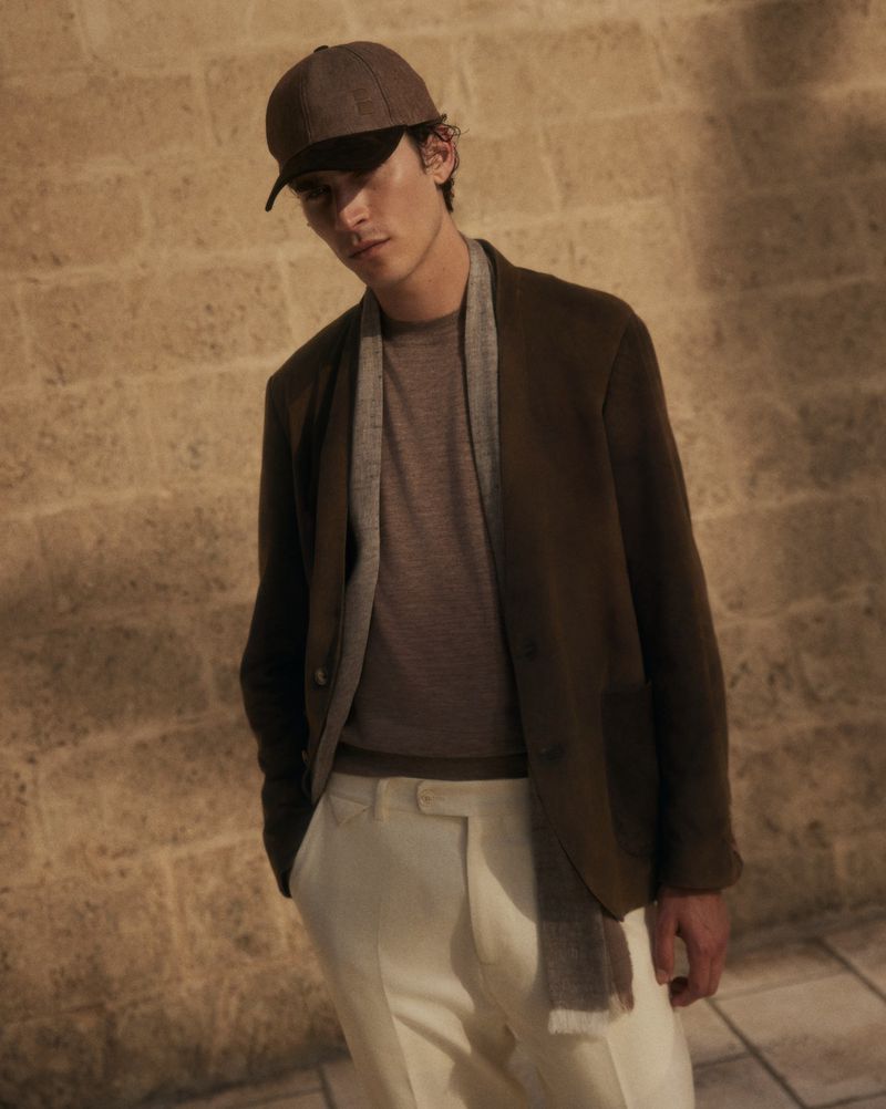 Masculine fashion: a brunette man in a blazer, t-shirt, baseball cap, and trousers poses against a textured wall in warm, soft lighting.