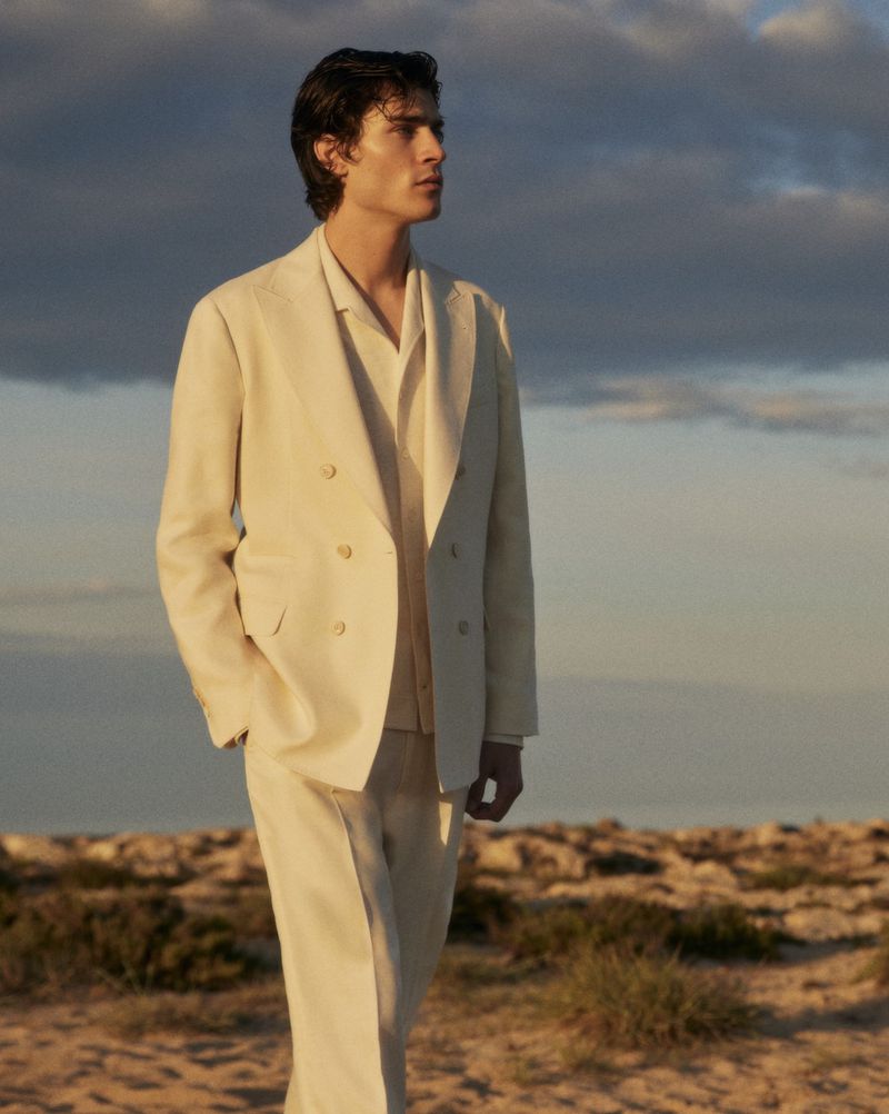Man in light-colored suit stands outdoors with sky background, facing right. Neutral tones and natural lighting give an editorial fashion look.
