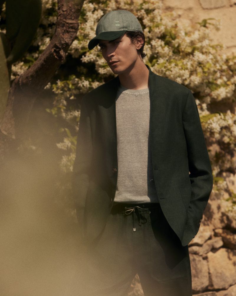 A man in a cap and green linen suit stands outdoors against a stone wall with flowering plants in soft, diffused daylight.