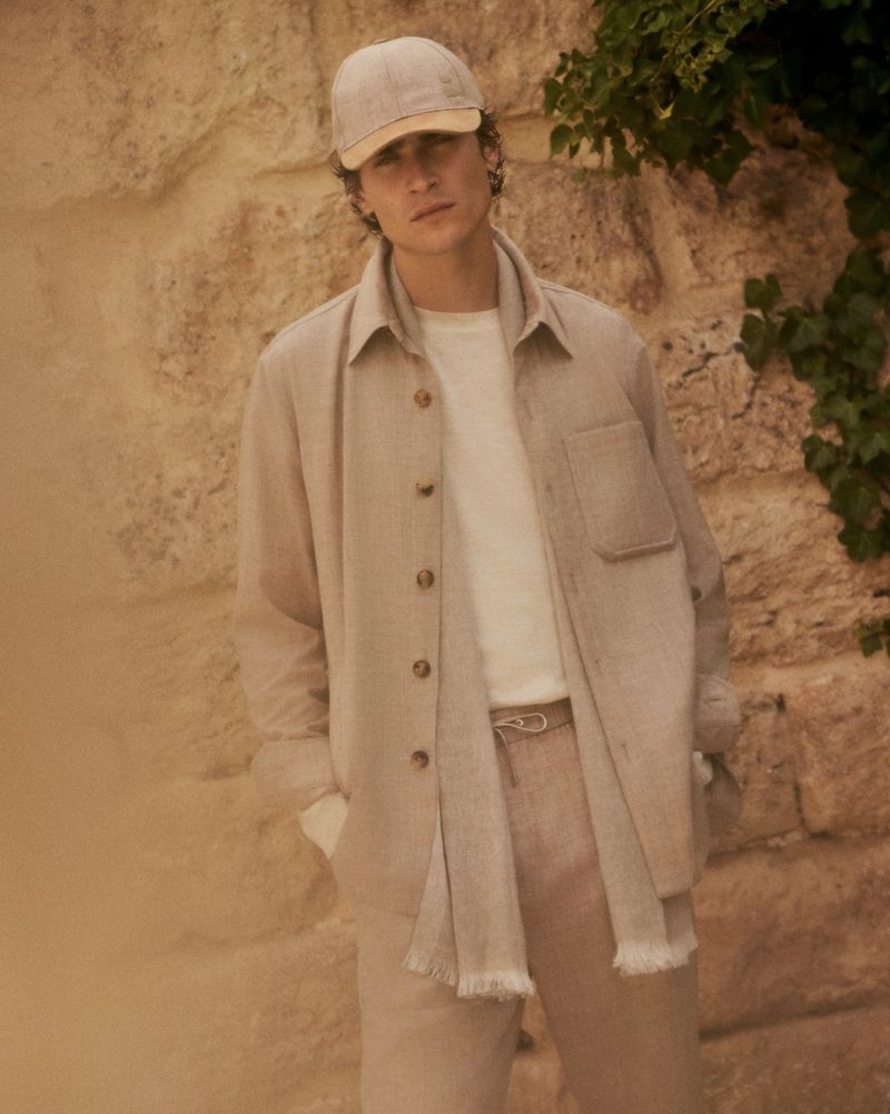 A stylish adult man wearing a light beige matching linen shirt and trousers, paired with a white t-shirt and baseball cap, poses outdoors.