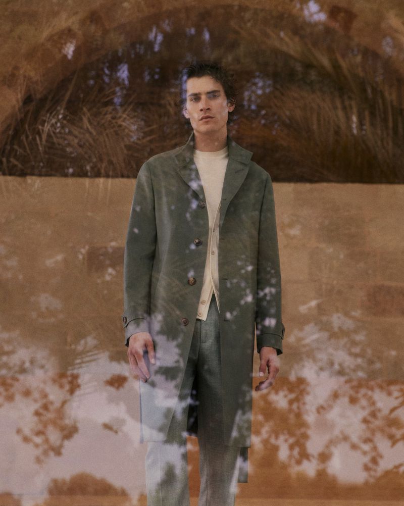 Man in olive green trench coat and pants against a textured wall with a tree branch shadow for fashion editorial, full shot.