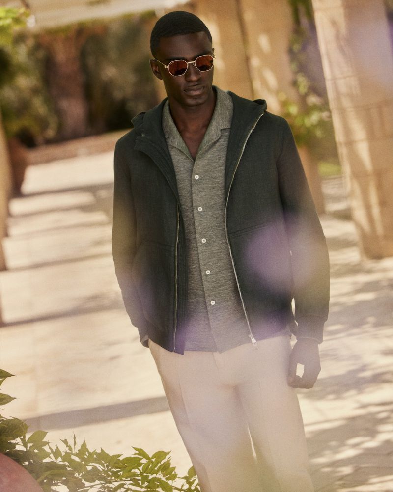 Portrait of a Black male model wearing sunglasses, a jacket, and beige pants, posing outdoors on a sunny day with architectural features.