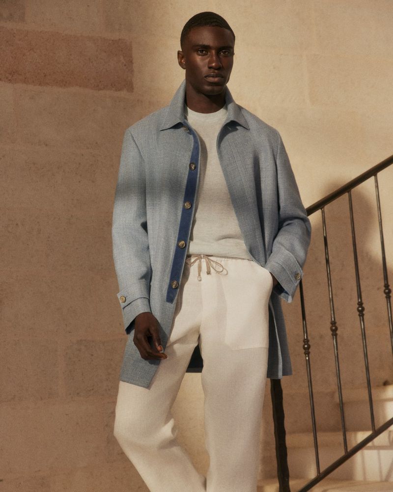 Man in blue coat, light sweater, and white linen pants poses on stairs. Indoor fashion shot with neutral tones, soft light, and minimalist style.