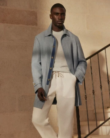 Man in blue coat, light sweater, and white linen pants poses on stairs. Indoor fashion shot with neutral tones, soft light, and minimalist style.