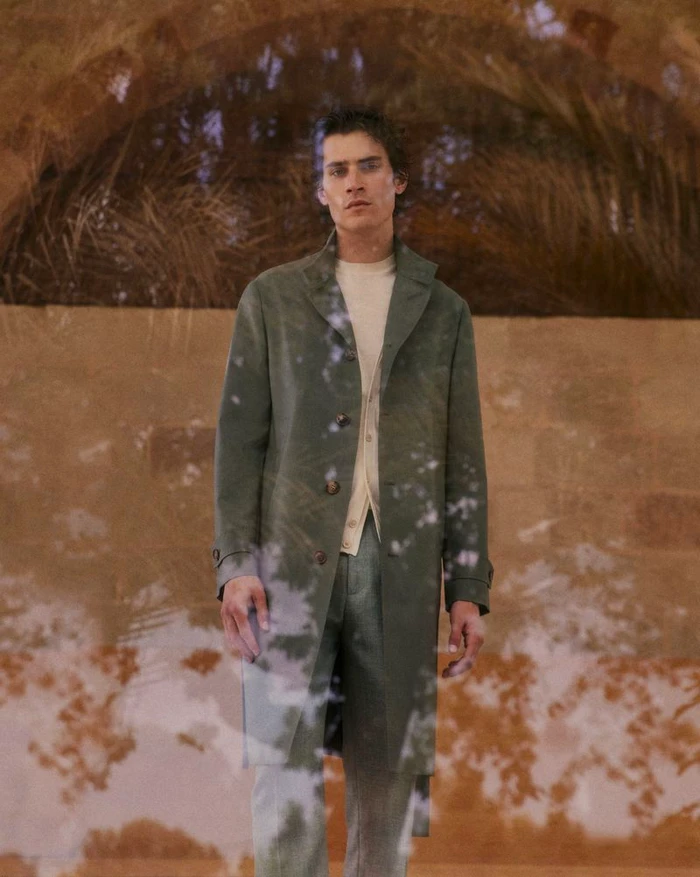 Model wearing a green coat and matching trousers over a cream shirt against a stone archway background