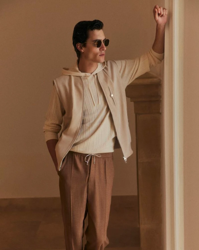 Model Liam Kelly wearing a cream hooded sweater, beige zip vest, and camel trousers leaning against an architectural stone arch.